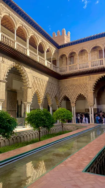 Cour intérieure de l’Alcázar de Séville avec arcades mudéjares richement sculptées, galeries à colonnes, orangers alignés et long bassin rectangulaire reflétant l’architecture mauresque sous un ciel bleu