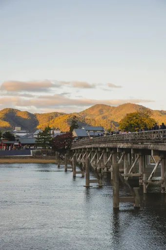 Picturesque view of old wooden Togetsukyo Bridge over calm river flowing near typical residential houses located at hillside against sunset sky in Kyoto