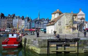 A bright scene of Honfleur’s harbor in Normandy, where historic, colorful houses line the old basin. In the foreground, a small red boat and the weathered stone quay enhance the authentic charm of the port. Visitors enjoy a sunny day in this picturesque maritime setting.