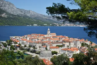 Panoramic view of Korčula, Croatia, with its fortified old town and red-tiled roofs standing out against the deep blue of the Adriatic Sea. Surrounded by mountains and pine trees, the historic town exudes an authentic and timeless Mediterranean charm.