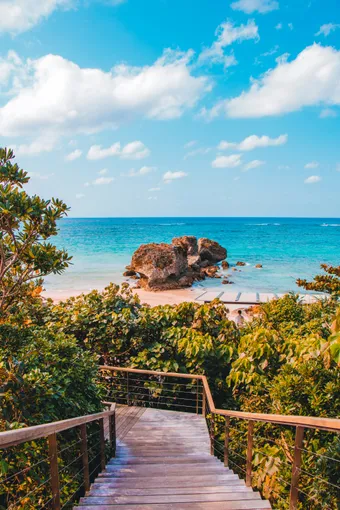A wooden staircase winds through lush tropical greenery and leads down to a pale sandy beach, bordered by the turquoise waters typical of Okinawa. In the distance, large rocks rise from the calm sea beneath a blue sky dotted with clouds, creating a peaceful, almost idyllic atmosphere.