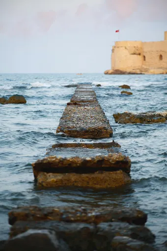 A stone jetty stretches out to a historic seaside castle under the bright sky.
