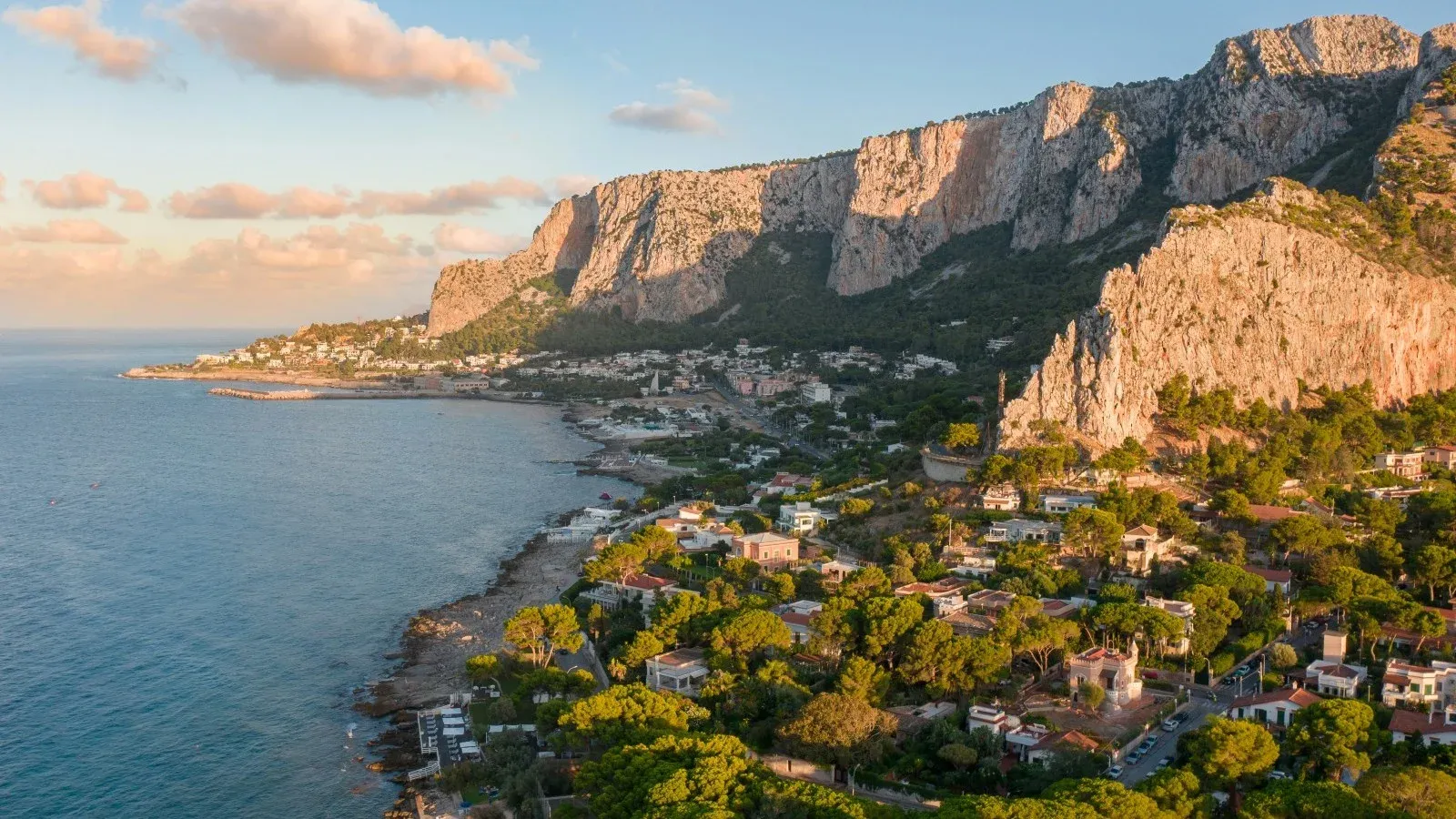Aerial view of a Sicilian coastal town nestled below limestone cliffs, bathed in warm sunset light.