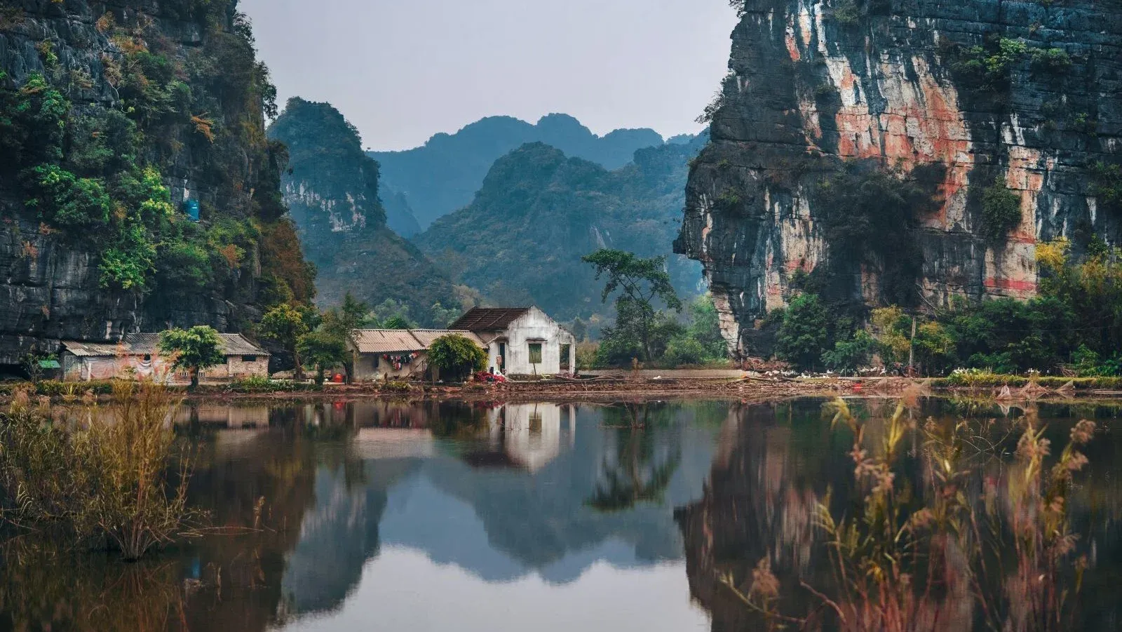 Peaceful landscape of Ninh Binh, Vietnam, featuring towering karst cliffs surrounding a traditional house by a calm body of water reflecting the mountains and vegetation.