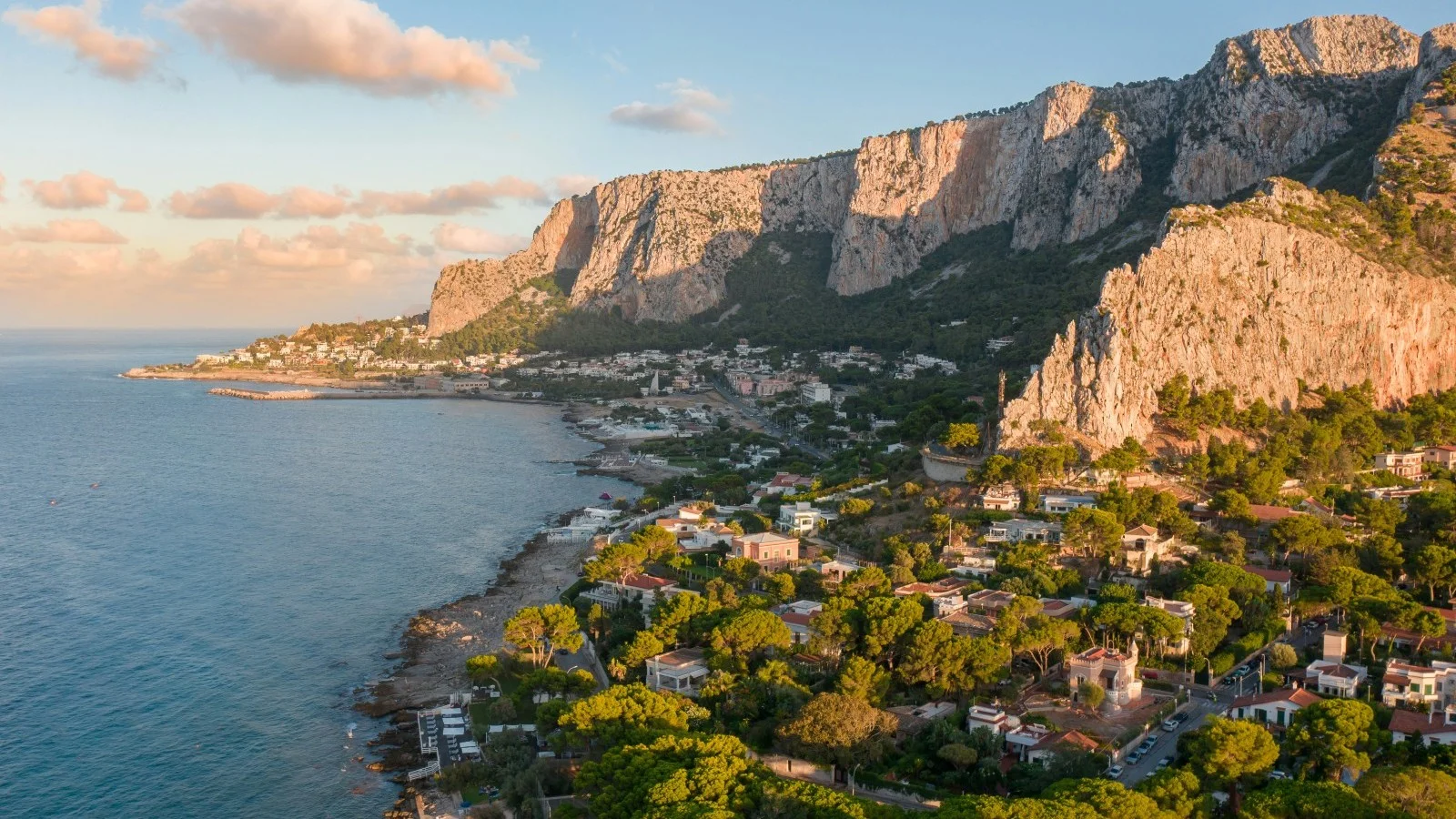 Vue aérienne d’un village côtier sicilien au pied de falaises calcaires, baigné par la lumière dorée du coucher de soleil.